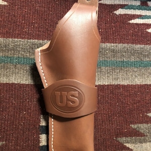 Op de afbeelding: Een bruine leren holster met witte stiksels en een US-logo dat op de voorkant is gegraveerd.