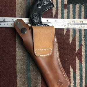 Op de afbeelding: Een bruine leren holster met een wit stikselontwerp. Een zwarte revolver met een adelaarontwerp zit in de holster.