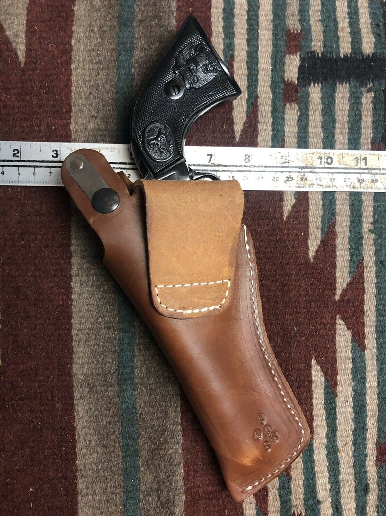 Op de afbeelding: Een bruine leren holster met een wit stikselontwerp. Een zwarte revolver met een adelaarontwerp zit in de holster.