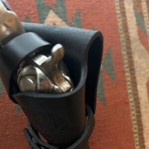May include: A black leather sheath with a silver object partially visible inside. The sheath has a strap with a buckle. The background is a patterned rug.