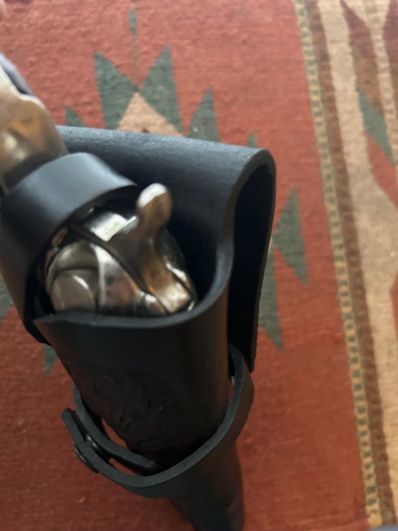 May include: A black leather sheath with a silver object partially visible inside. The sheath has a strap with a buckle. The background is a patterned rug.