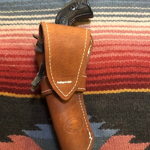 May include: A brown leather holster with white stitching and a black handgun inside. The holster has a maker's mark that reads "Handcrafted by Maverick".