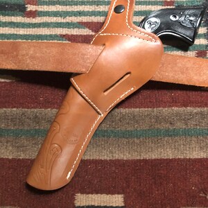May include: A brown leather holster with white stitching and a black gun inside. The holster is on a brown and green patterned rug.