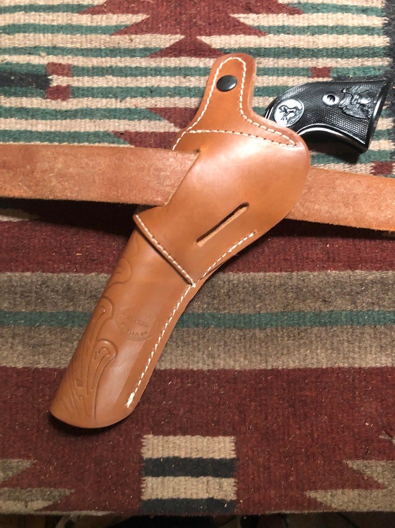 May include: A brown leather holster with white stitching and a black gun inside. The holster is on a brown and green patterned rug.