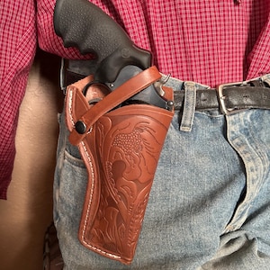 May include: A brown leather holster with a floral design, holding a black revolver. The holster is attached to a belt worn with blue denim jeans and a red checked shirt.