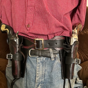 Puede incluir: Una persona que lleva una camisa de cuadros rojos y pantalones vaqueros azules con un cinturón de cuero marrón y dos fundas de cuero negro con hebillas plateadas, cada una con un revólver plateado.
