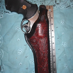 Puede incluir: Un revólver plateado con mango negro en una funda de cuero marrón con un intrincado diseño floral. La funda tiene una solapa que cubre la protección del gatillo. Una regla es visible en el lado derecho de la imagen.