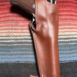 May include: A brown leather holster with a stitched seam down the side. A black handgun is inside the holster.
