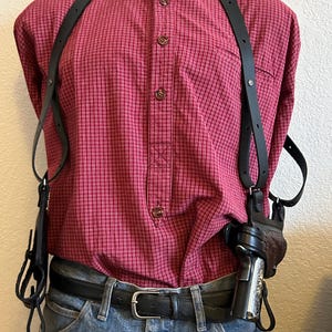Puede incluir: Un holster de cuero negro con dos correas que pasan por encima de los hombros. El holster se lleva sobre una camisa de cuadros rojos y blancos y unos vaqueros azules. Hay una pistola en el holster.
