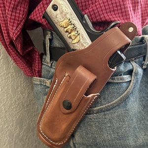 May include: A brown leather holster with a snap button fastening, holding a silver and black handgun with gold accents. The holster is worn on a person's belt.