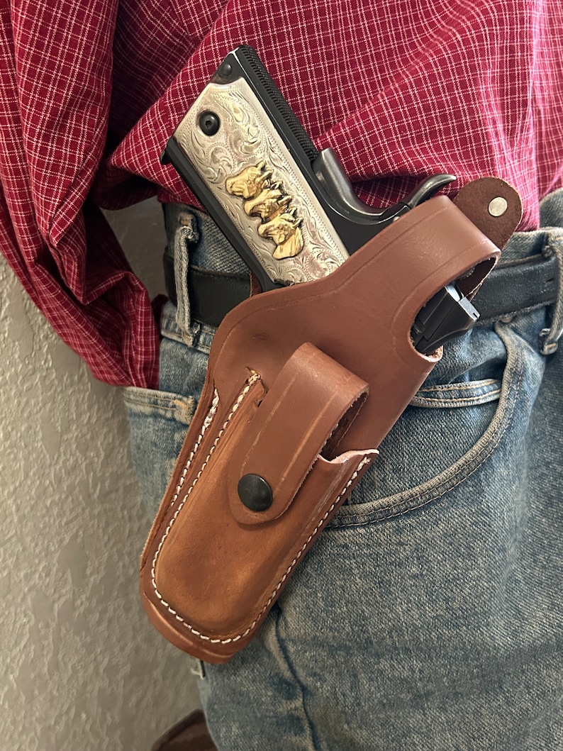 May include: A brown leather holster with a snap button fastening, holding a silver and black handgun with gold accents. The holster is worn on a person's belt.