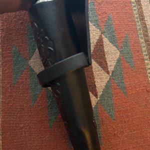 May include: A black leather bottle holder with a decorative floral pattern. The holder has a loop and a protective flap. The holder is designed to carry a bottle. The holder is shown against a patterned rug.