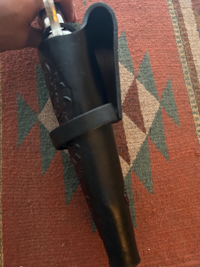 May include: A black leather bottle holder with a decorative floral pattern. The holder has a loop and a protective flap. The holder is designed to carry a bottle. The holder is shown against a patterned rug.