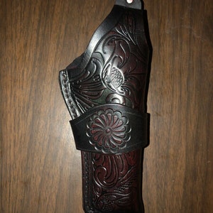 May include: A black leather holster with intricate floral designs embossed on the surface. The holster has a brown leather strap with a decorative flower design.
