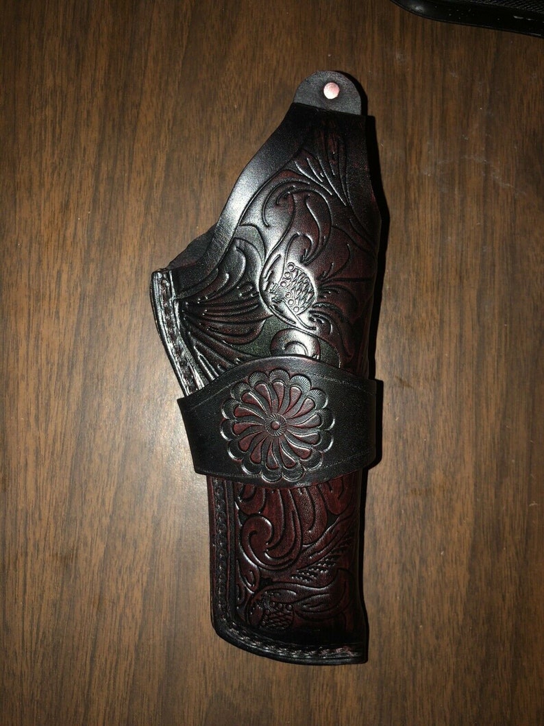 May include: A black leather holster with intricate floral designs embossed on the surface. The holster has a brown leather strap with a decorative flower design.