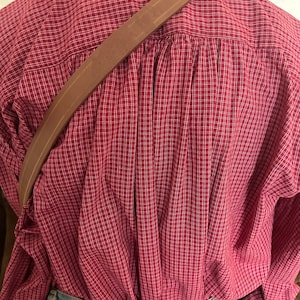 Pode incluir: Uma al&ccedil;a de ombro de couro marrom com uma fivela. A al&ccedil;a &eacute; usada sobre uma camisa xadrez vermelha e branca.