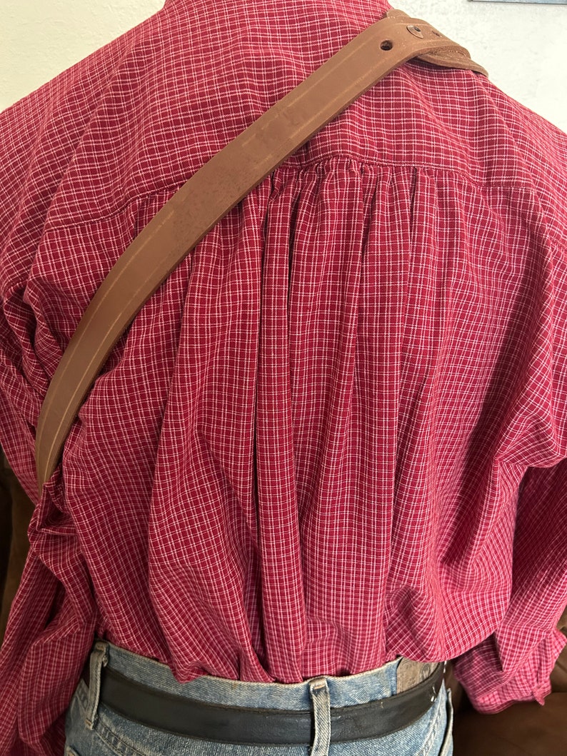 Pode incluir: Uma al&ccedil;a de ombro de couro marrom com uma fivela. A al&ccedil;a &eacute; usada sobre uma camisa xadrez vermelha e branca.