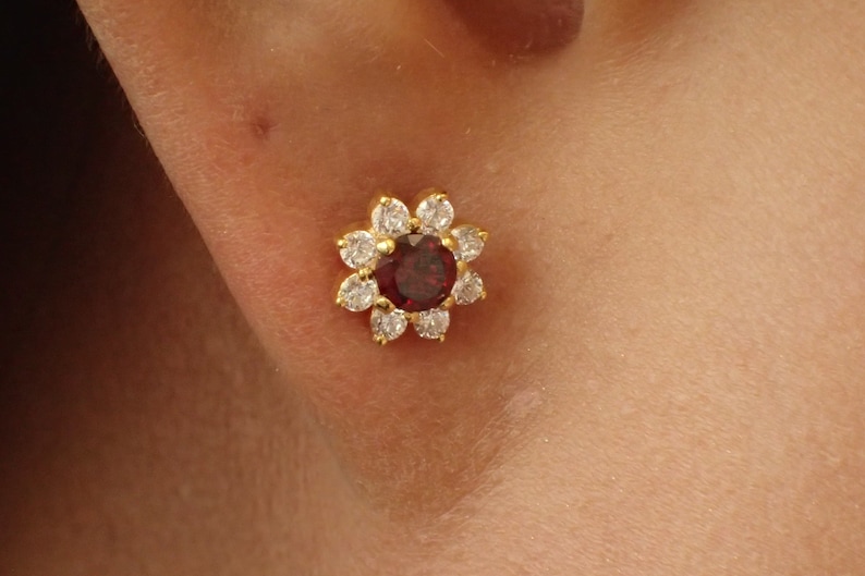 K&ouml;nnte beinhalten: Ein goldener, blumenf&ouml;rmiger Ohrring mit einem roten Edelstein in der Mitte und klaren Zirkonia-Steinen am Rand.