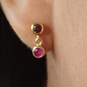 May include: Gold dangle earrings with two red gemstones. The top gemstone is a darker red and the bottom gemstone is a lighter pink.