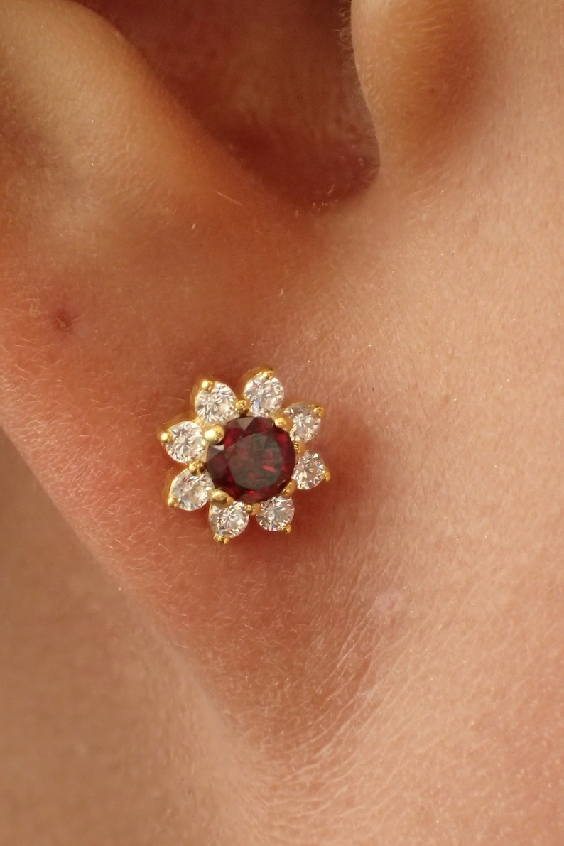 K&ouml;nnte beinhalten: Ein goldener Ohrring in Blumenform mit einem roten Edelstein in der Mitte und klaren Zirkonia-Steinen am Rand.