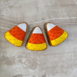 Può includere: Tre decorazioni in feltro a forma di caramelle di mais. Ogni caramella di mais è gialla, arancione e bianca, con un bordo marrone e piccoli accenti di perline. I pezzi di feltro sono disposti su una superficie chiara.