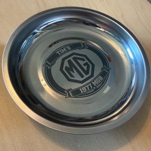 May include: A shiny, stainless steel bowl with a polished finish. The bowl's interior features a dark gray emblem with the letters "MG" inside a shield, along with the text "TIM'S" and "1977 MGB".