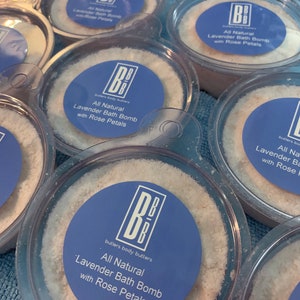 Puede incluir: Se muestran varias bombas de baño de lavanda totalmente naturales con pétalos de rosa en recipientes de plástico transparente. Cada recipiente tiene una etiqueta azul con el texto "All Natural Lavender Bath Bomb with Rose Petals". Las bombas de baño son blancas y granulares.