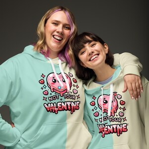Könnte beinhalten: Zwei Personen tragen passende Kapuzenpullover. Die Kapuzenpullover sind vertikal geteilt, eine Seite mintgrün und die andere cremefarben. Jeder Kapuzenpullover zeigt eine Cartoon-Herz-Grafik mit dem Text "NOT YOUR VALENTINE" in Rot.