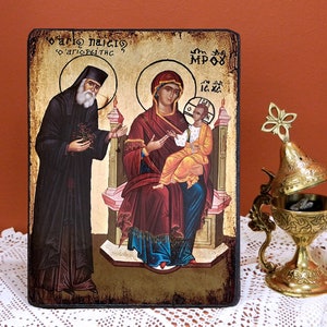Könnte beinhalten: Eine handgemalte Ikone, die die Jungfrau Maria und das Kind darstellt, mit dem Heiligen Paisios vom Berg Athos, der links steht. Die Ikone ist in einem dunklen Holzrahmen gerahmt und hat einen goldenen Hintergrund. Der Text auf der Ikone lautet "O' Agios Paisios, o' Apostolos tis", was übersetzt "Heiliger Paisios, der Apostel der" bedeutet.