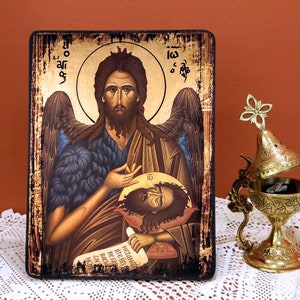 Puede incluir: Un icono pintado a mano de Juan el Bautista sosteniendo la cabeza de Juan el Bautista. El icono está en un panel de madera con un fondo dorado y un marco negro. El icono está rodeado por un halo y alas. El icono es una obra de arte religiosa.
