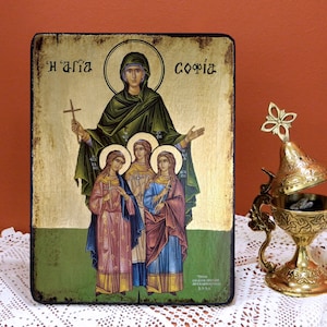 May include: A wooden icon depicting Saint Sophia and her three daughters, Faith, Hope, and Charity. The icon is painted in a traditional style with gold accents and a green background. The text "Η' Αγία Σοφία" is written in Greek at the top of the icon.