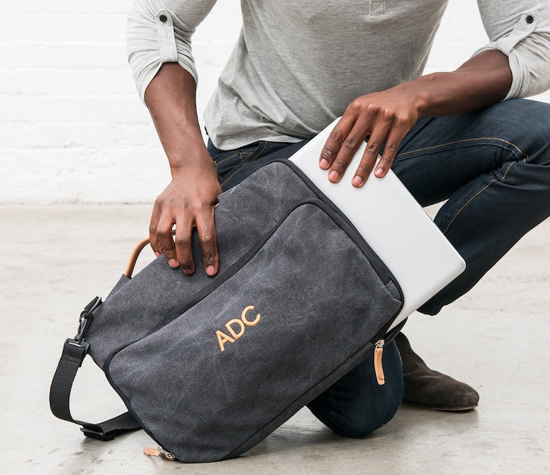 May include: A black canvas laptop bag with a brown leather strap and the initials "ADC" embroidered in gold thread.