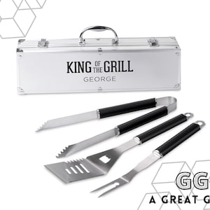 May include: A silver grilling tool set with black handles and a matching case. The case is engraved with "KING OF THE GRILL" and a name. The set includes a spatula, fork, tongs, and knife. The text "A GREAT GIFT!" is also visible.