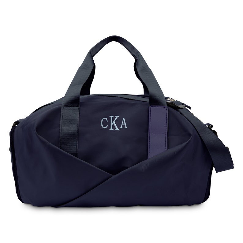 May include: A navy blue duffel bag with a black zipper and a black strap. The bag has a leather patch on the side with the initials "CKA" embroidered in white.