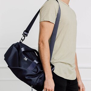 May include: A black duffel bag with a navy blue strap and a white "M" embroidered on the front. The bag is being carried by a person wearing a light green t-shirt and black pants.
