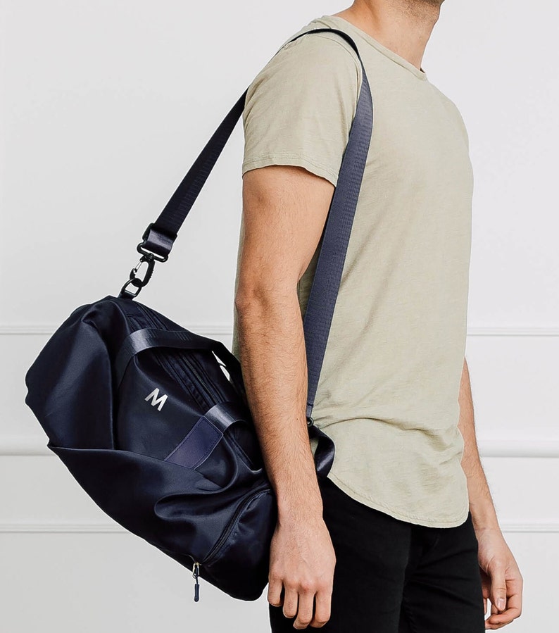 May include: A black duffel bag with a navy blue strap and a white "M" embroidered on the front. The bag is being carried by a person wearing a light green t-shirt and black pants.
