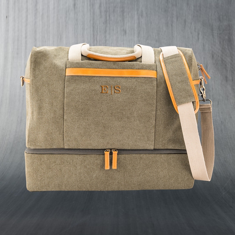 May include: Olive green canvas duffel bag with tan accents and a detachable shoulder strap. Features a front pocket with the initials "ES" in a brown color. Includes a separate bottom compartment and tan handles.