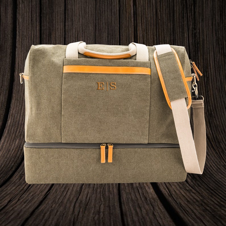 May include: Olive green canvas duffel bag with tan leather accents and a detachable shoulder strap. Features a front pocket with the embroidered initials "E|S". Includes a separate bottom compartment and tan handles.