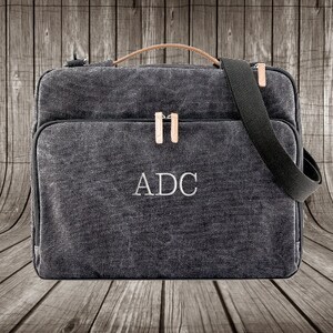 May include: A dark gray canvas messenger bag with a leather handle and a black adjustable strap. The bag has a zipper closure and a front pocket. The initials "ADC" are embroidered on the front of the bag.