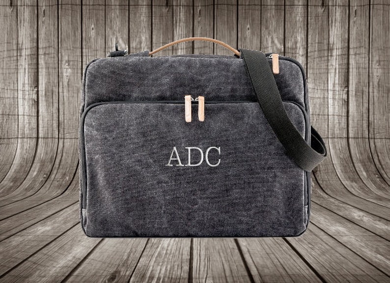 May include: A dark gray canvas messenger bag with a leather handle and a black adjustable strap. The bag has a zipper closure and a front pocket. The initials "ADC" are embroidered on the front of the bag.