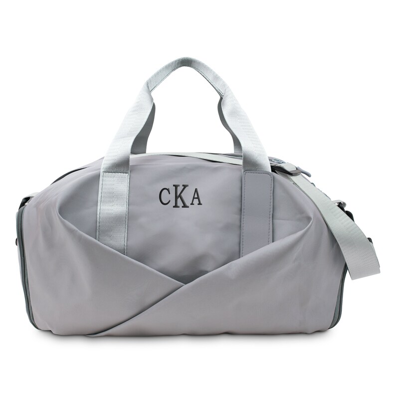 Op de afbeelding: Een grijze reistas met een ritssluiting en een bovenste handvat. De tas heeft een witte band en de initialen "CKA" zijn geborduurd op de voorkant.