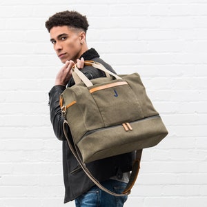May include: Olive green canvas duffel bag with tan accents and a navy blue "J" initial. The bag features a top handle, a shoulder strap, and a bottom compartment. The bag is approximately 20 inches long.