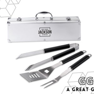 Peut inclure: Un ensemble d'ustensiles de barbecue en métal argenté avec des manches noirs, présenté dans une mallette argentée. La mallette est gravée de "BEST MAN JACKSON 07.09.22". L'ensemble comprend une spatule, une fourchette, des pinces et un couteau.