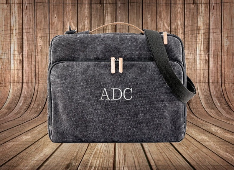 May include: A black canvas laptop bag with a leather handle and a shoulder strap. The bag has a zipper closure and the initials "ADC" are embroidered on the front.