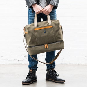 May include: Olive green canvas duffel bag with tan leather accents and a blue embroidered "J". The bag has a separate bottom compartment, a shoulder strap, and tan handles. The bag is held by a person wearing blue jeans and black boots.