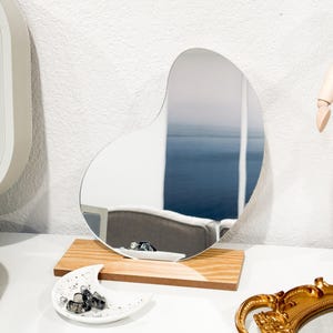 May include: A wooden stand with a unique, freeform shaped mirror. The mirror is reflecting a room with a chair and a window. The stand is on a white surface with a white ceramic crescent moon dish with black stones.