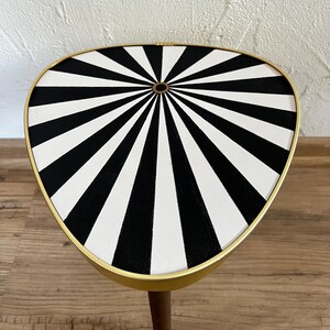 May include: A triangular side table with a gold-toned metal rim and a flat top surface featuring a black and white radial sunburst pattern. The table has a single visible wooden leg extending downward from the center of the underside.