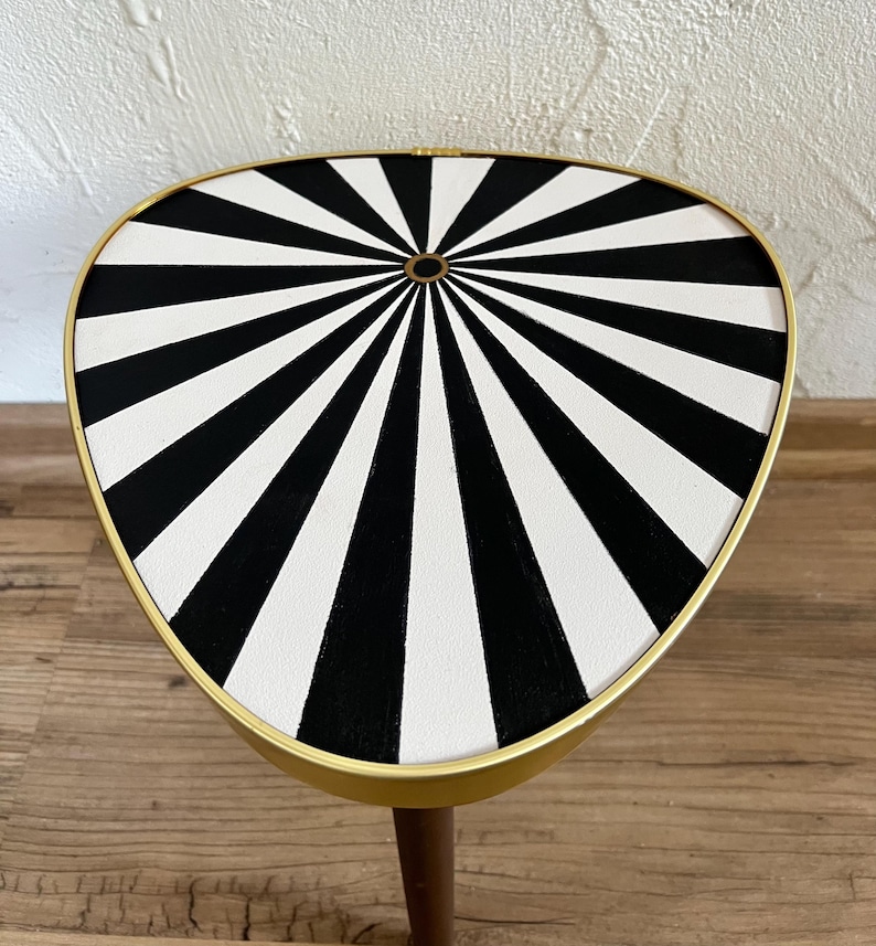May include: A triangular side table with a gold-toned metal rim and a flat top surface featuring a black and white radial sunburst pattern. The table has a single visible wooden leg extending downward from the center of the underside.