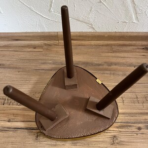 May include: A three-legged stool viewed from the underside, featuring a triangular seat with rounded corners and a metallic gold-toned edge. The three dark brown, cylindrical legs are attached to the seat with rectangular wooden mounts.