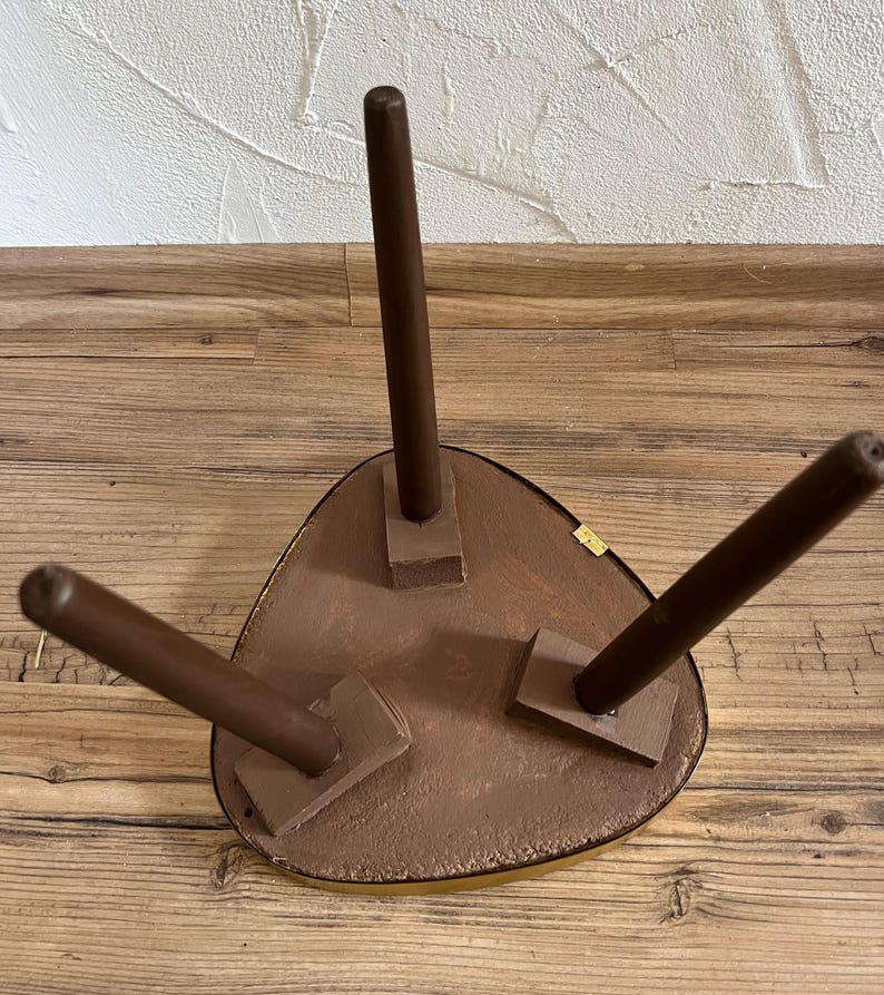 May include: A three-legged stool viewed from the underside, featuring a triangular seat with rounded corners and a metallic gold-toned edge. The three dark brown, cylindrical legs are attached to the seat with rectangular wooden mounts.
